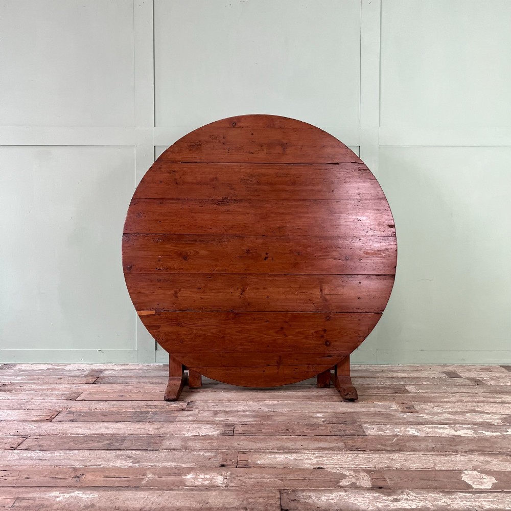 A 19th-century French circular tilt-top vendange table constructed from warm brown planked timber, shown tilted upright against a pale green wall.