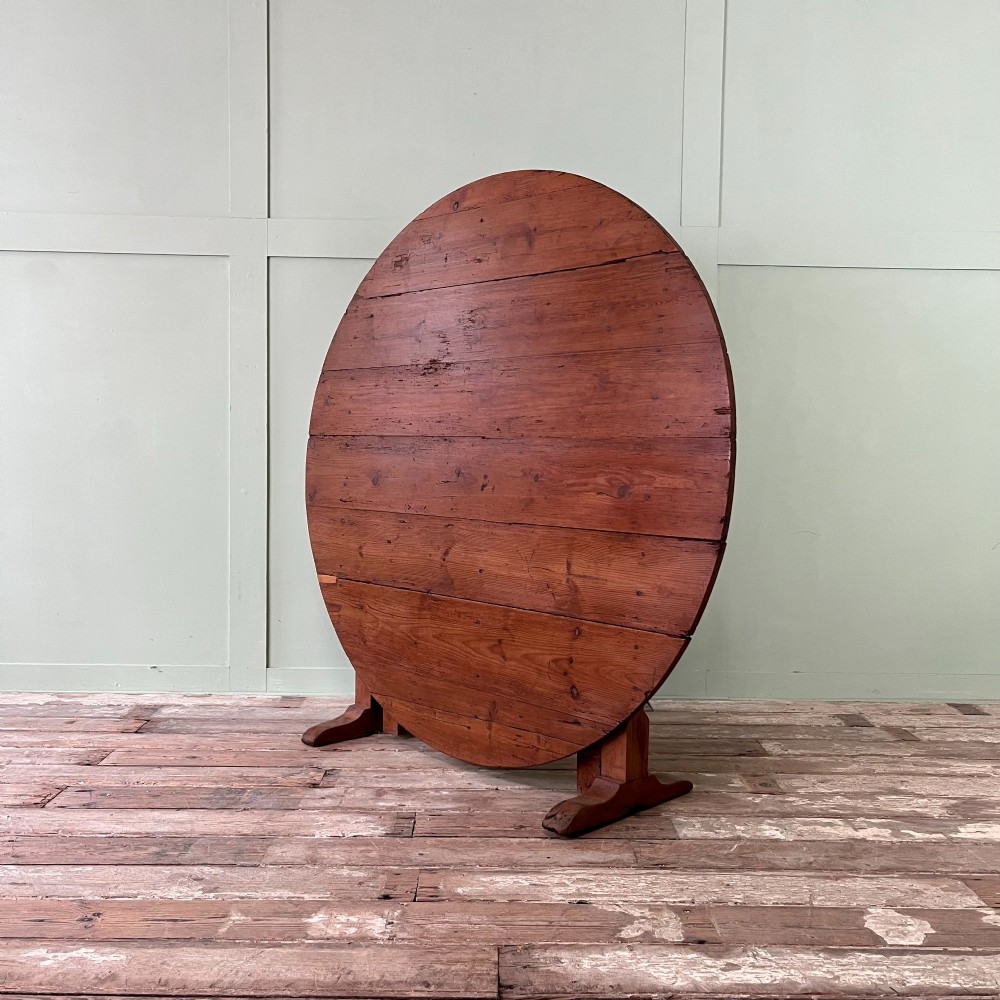 A 19th-century French circular tilt-top vendange table constructed from warm brown planked timber, shown tilted upright against a pale green wall. View IV