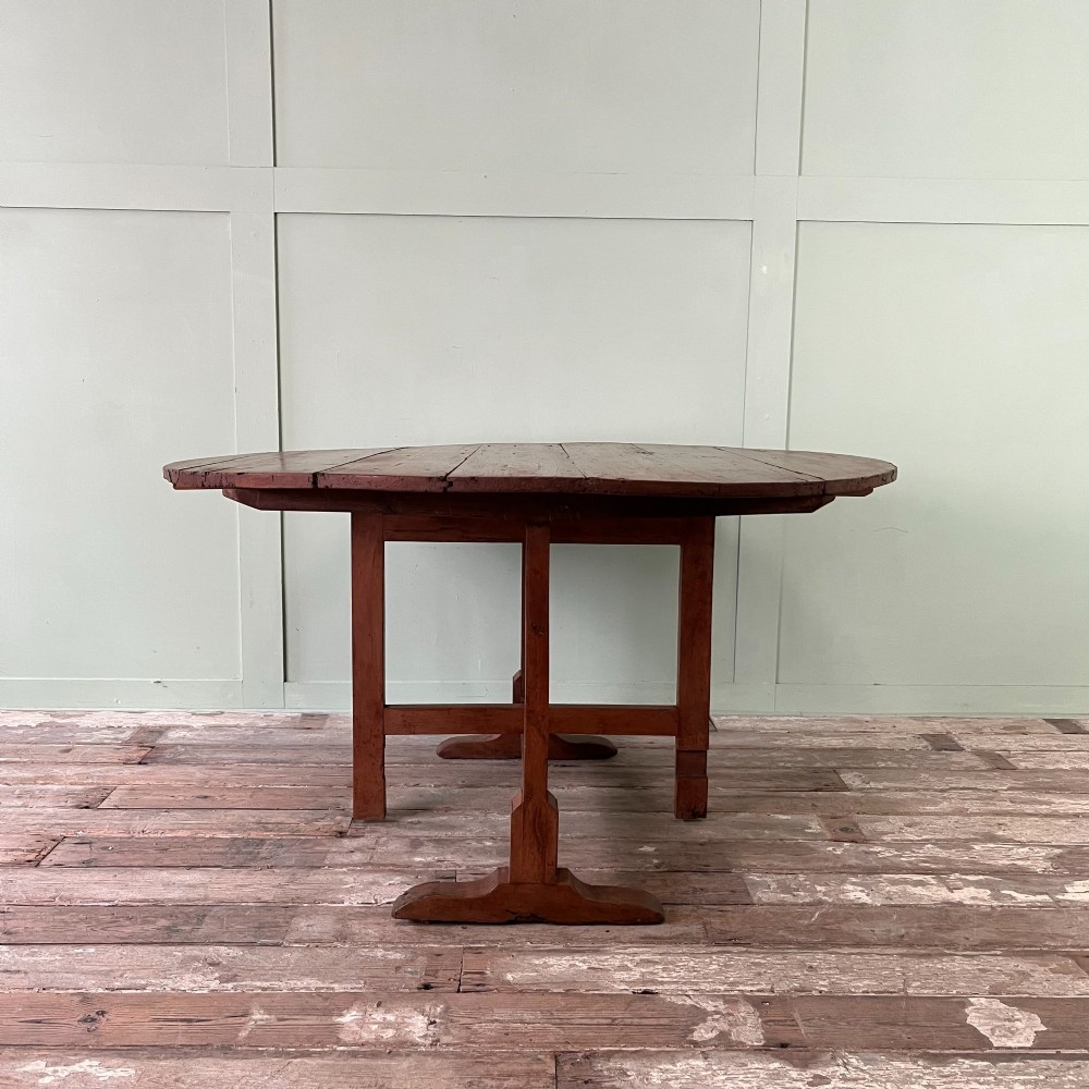 A 19th-century French circular tilt-top vendange table constructed from warm brown planked timber, shown tilted upright against a pale green wall. View IX