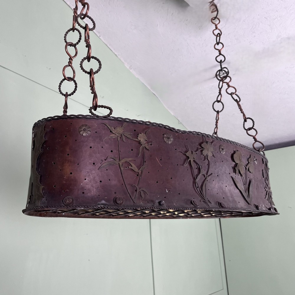 An oval Arts and Crafts copper pendant light featuring hand-chased floral motifs suspended from twin copper link chains with ceiling roses. View V