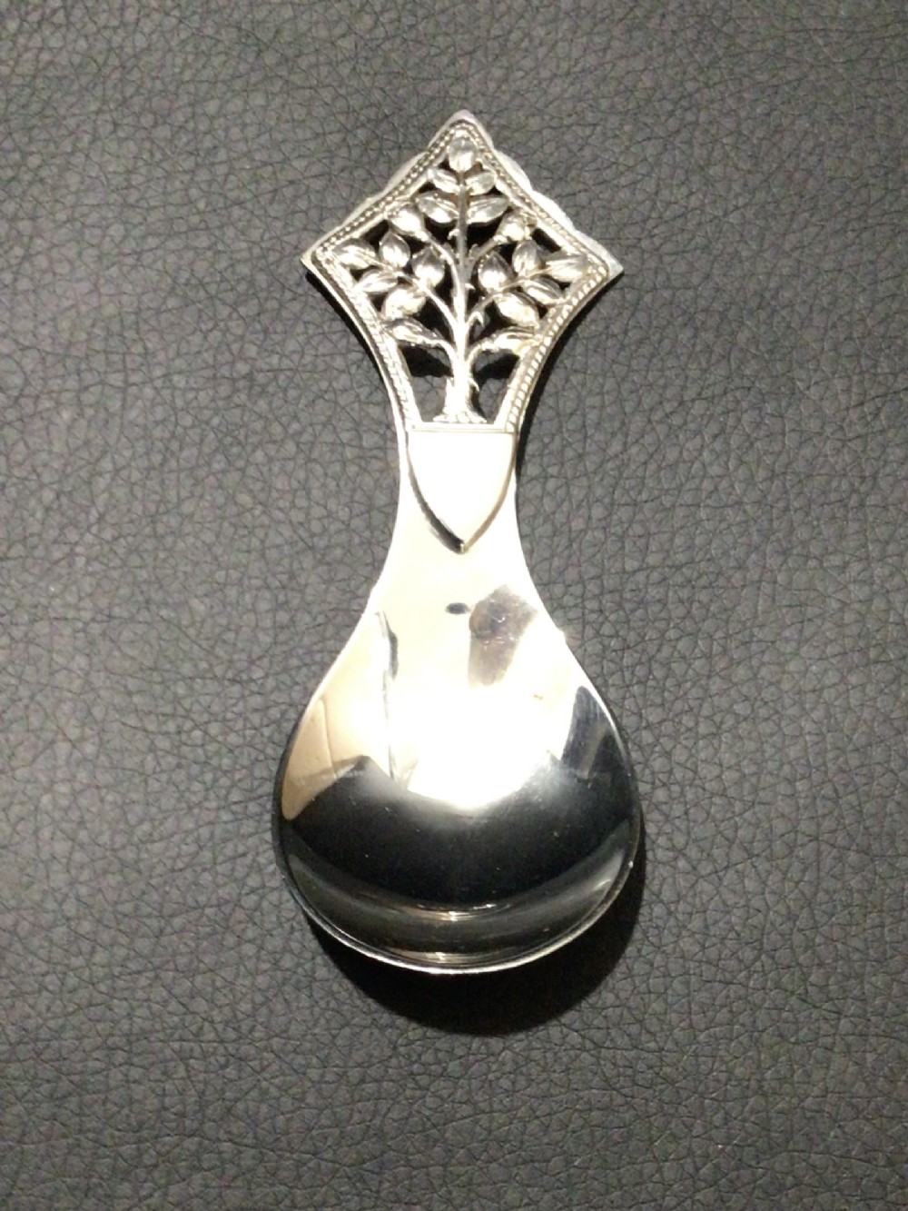 A sterling silver tea caddy spoon featuring a pierced diamond-shaped handle decorated with a tree of life motif and a polished pear-shaped bowl.