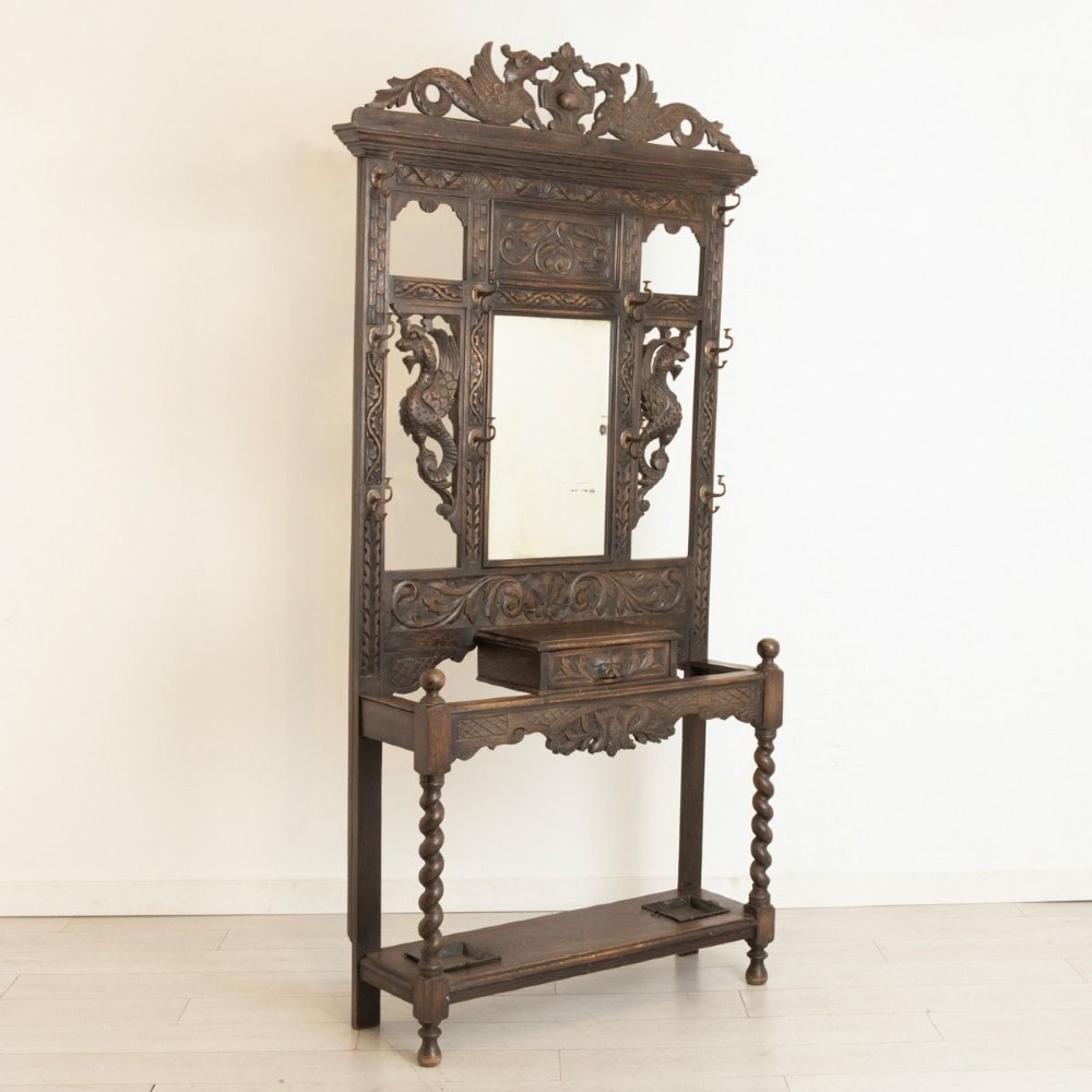 A late Victorian carved dark oak hall stand featuring a central mirror, dragon motifs, barley twist supports, integrated coat hooks, and an umbrella stand base.