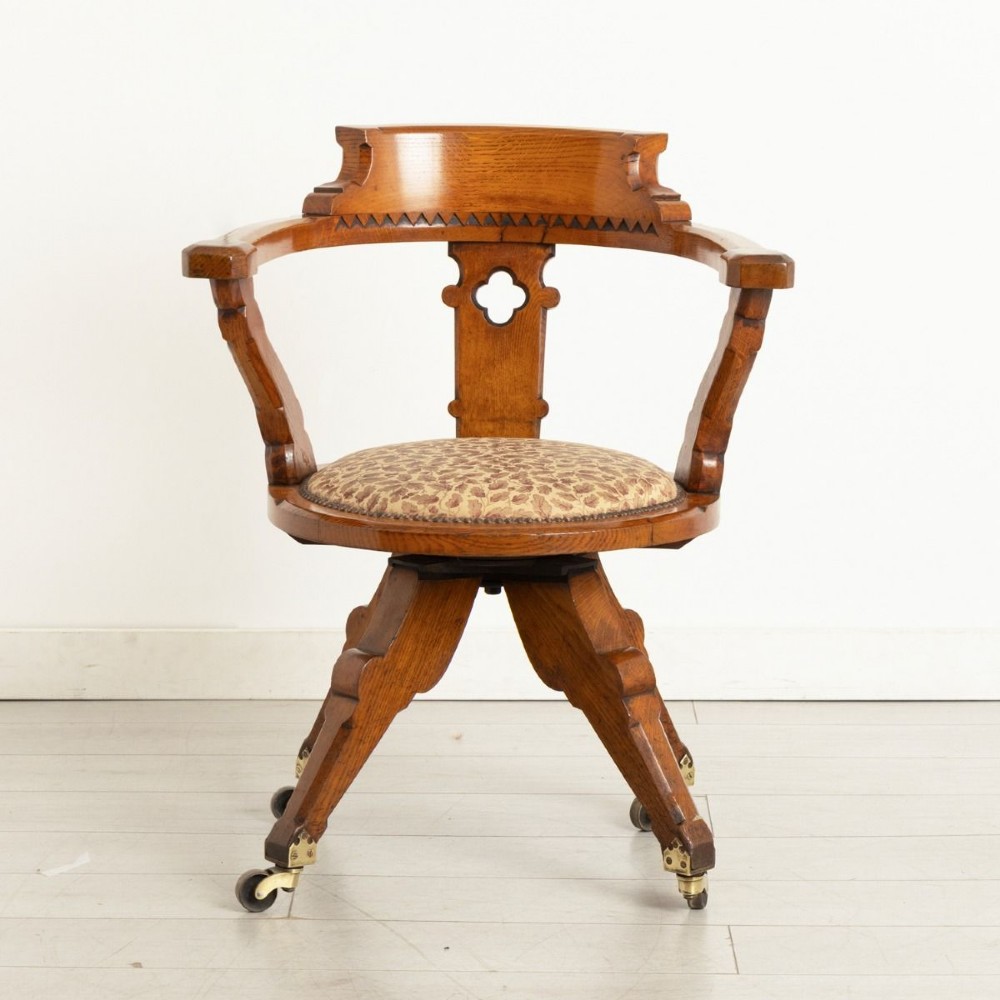 A Victorian Gothic Revival oak swivel desk chair featuring a curved crest rail, pierced quatrefoil splat back, upholstered seat, and splayed legs on brass castors.