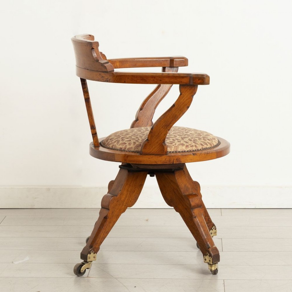 A Victorian Gothic Revival oak swivel desk chair featuring a curved crest rail, pierced quatrefoil splat back, upholstered seat, and splayed legs on brass castors. View II