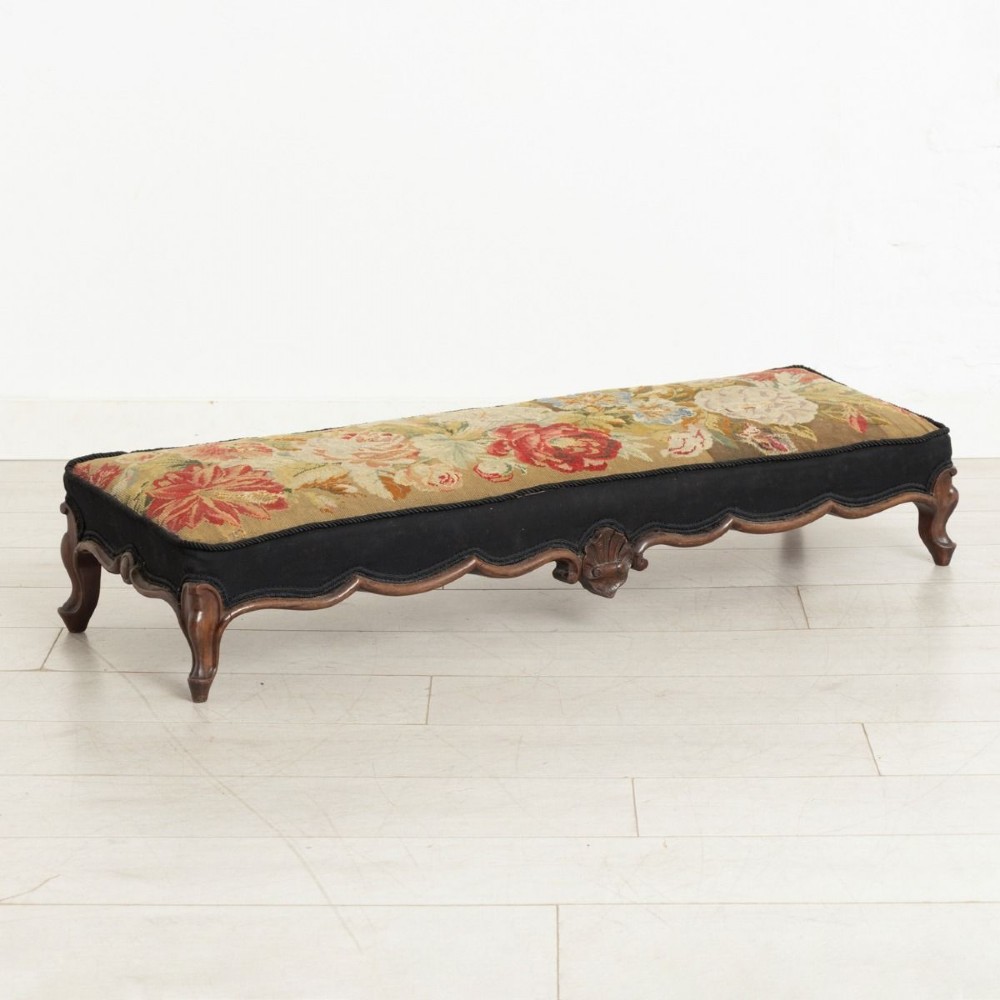 A low rectangular Victorian footstool crafted from carved dark walnut with short cabriole legs and floral tapestry upholstery.