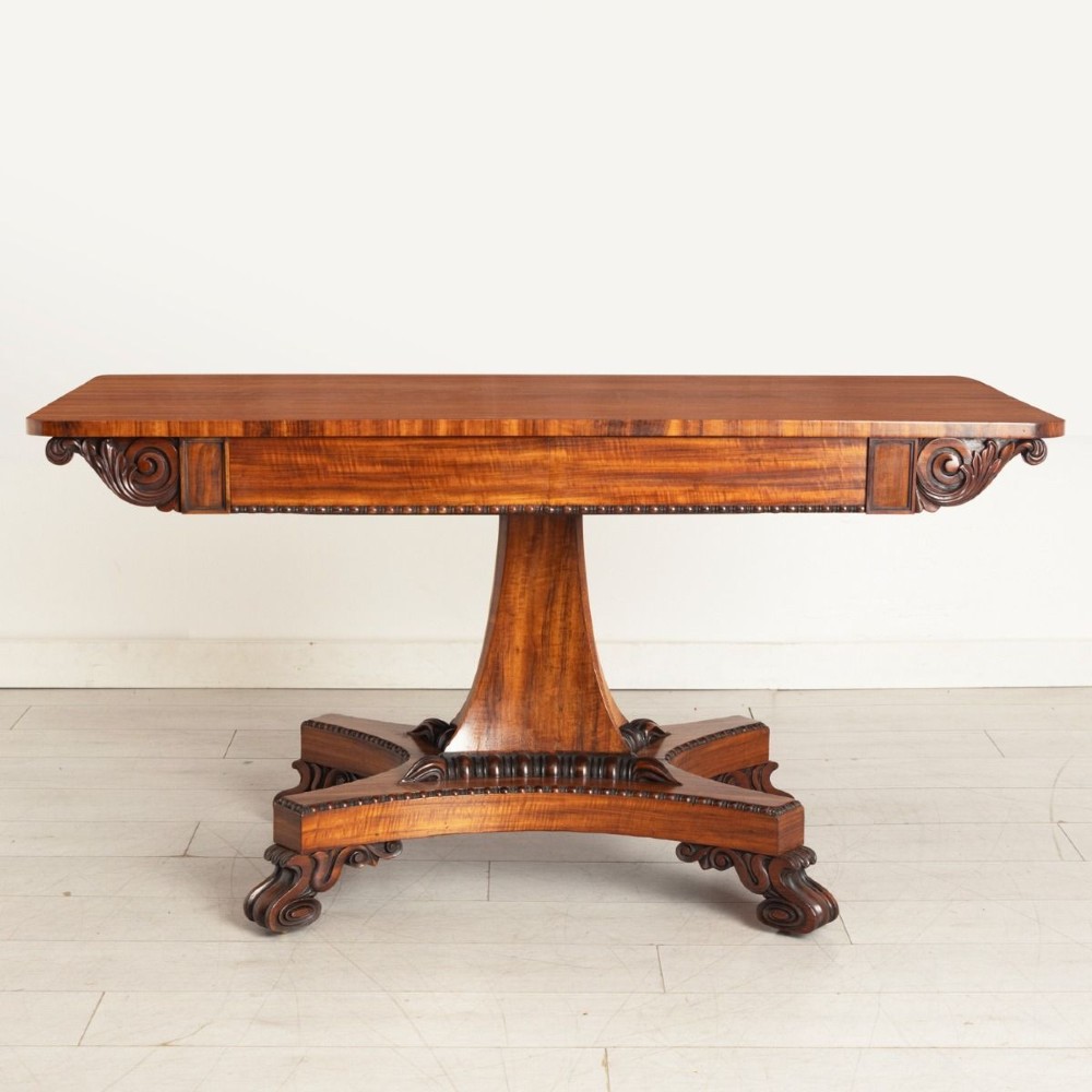 A Regency period Goncalo Alves library table featuring a rectangular top, frieze with carved brackets, a central flared pedestal support, and a quadform base with scroll feet. View II