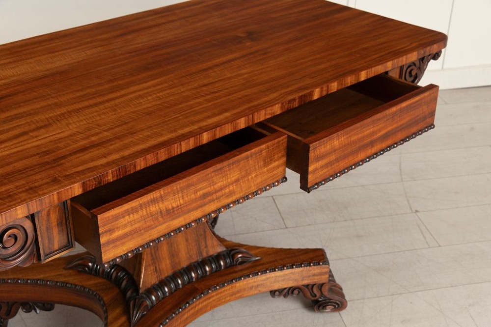 A Regency period Goncalo Alves library table featuring a rectangular top, frieze with carved brackets, a central flared pedestal support, and a quadform base with scroll feet. View III
