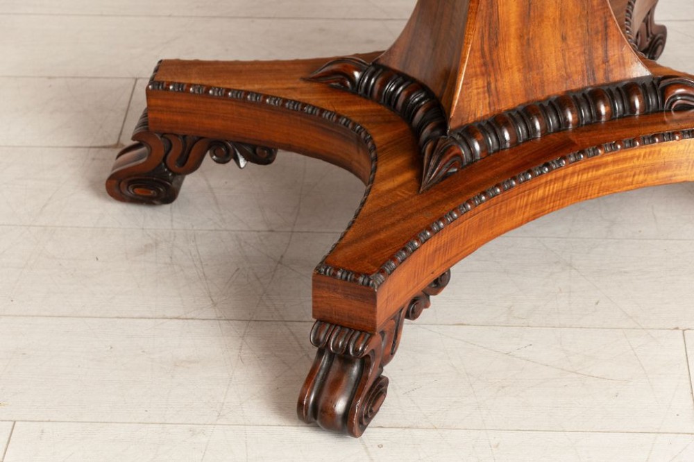 A Regency period Goncalo Alves library table featuring a rectangular top, frieze with carved brackets, a central flared pedestal support, and a quadform base with scroll feet. View IV