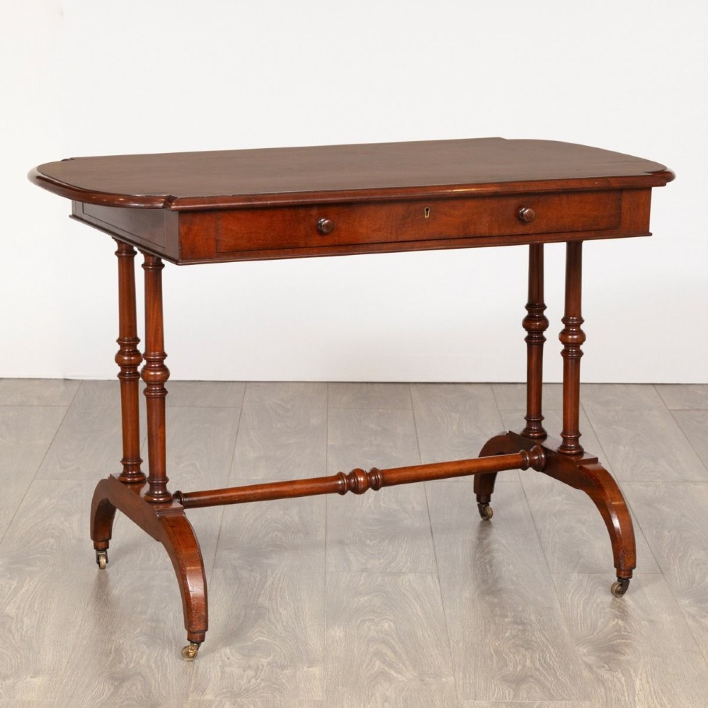 A Regency mahogany writing table featuring a rectangular top with rounded corners, a single frieze drawer with turned wooden knobs, twin turned end supports, a central turned stretcher, and splayed legs resting on brass castors.