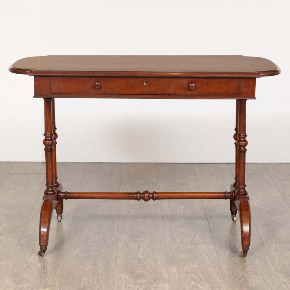 A Regency mahogany writing table featuring a rectangular top with rounded corners, a single frieze drawer with turned wooden knobs, twin turned end supports, a central turned stretcher, and splayed legs resting on brass castors. View II