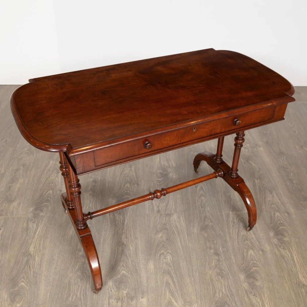 A Regency mahogany writing table featuring a rectangular top with rounded corners, a single frieze drawer with turned wooden knobs, twin turned end supports, a central turned stretcher, and splayed legs resting on brass castors. View III