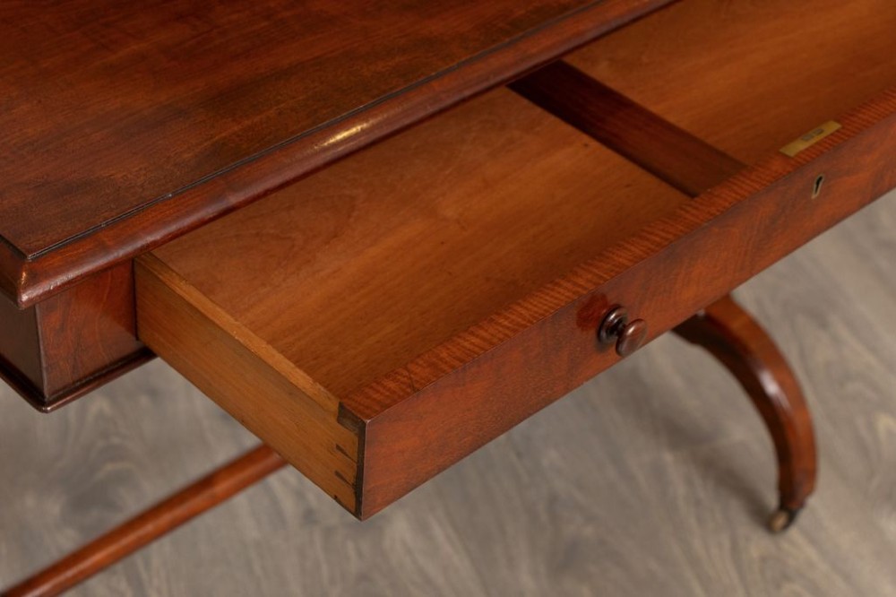 A Regency mahogany writing table featuring a rectangular top with rounded corners, a single frieze drawer with turned wooden knobs, twin turned end supports, a central turned stretcher, and splayed legs resting on brass castors. View IV