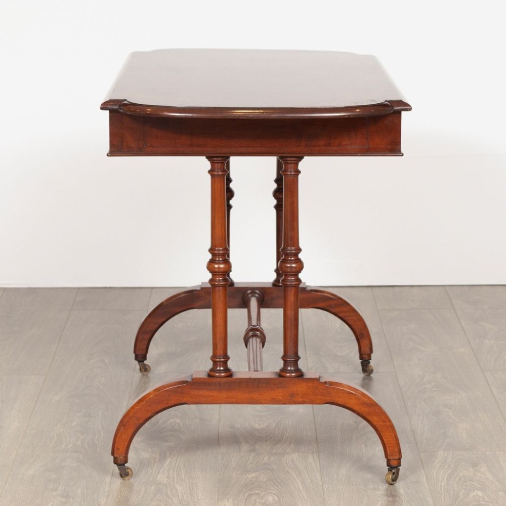 A Regency mahogany writing table featuring a rectangular top with rounded corners, a single frieze drawer with turned wooden knobs, twin turned end supports, a central turned stretcher, and splayed legs resting on brass castors. View V
