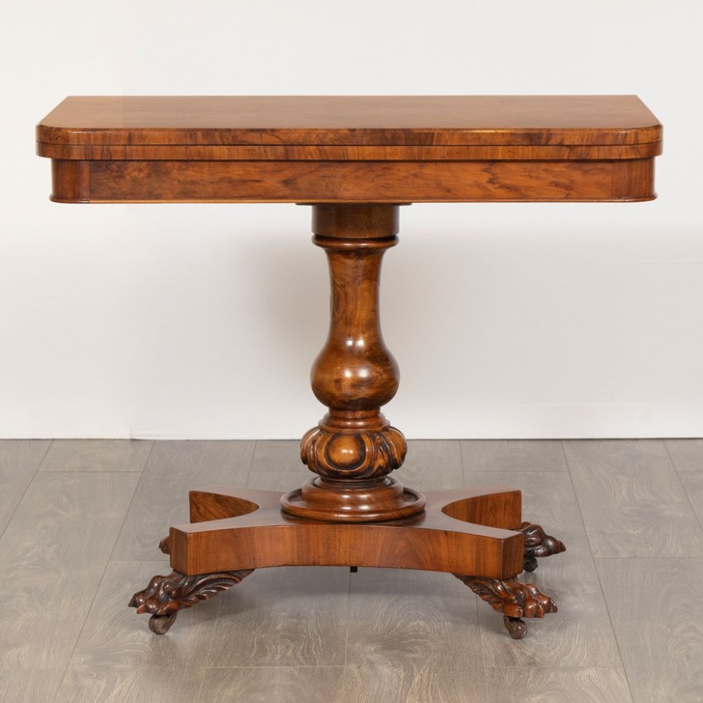 A mid-19th century Victorian burr walnut games table featuring a fold-over rectangular top resting on a turned bulbous pedestal column and a quadripartite base with carved feet on castors.