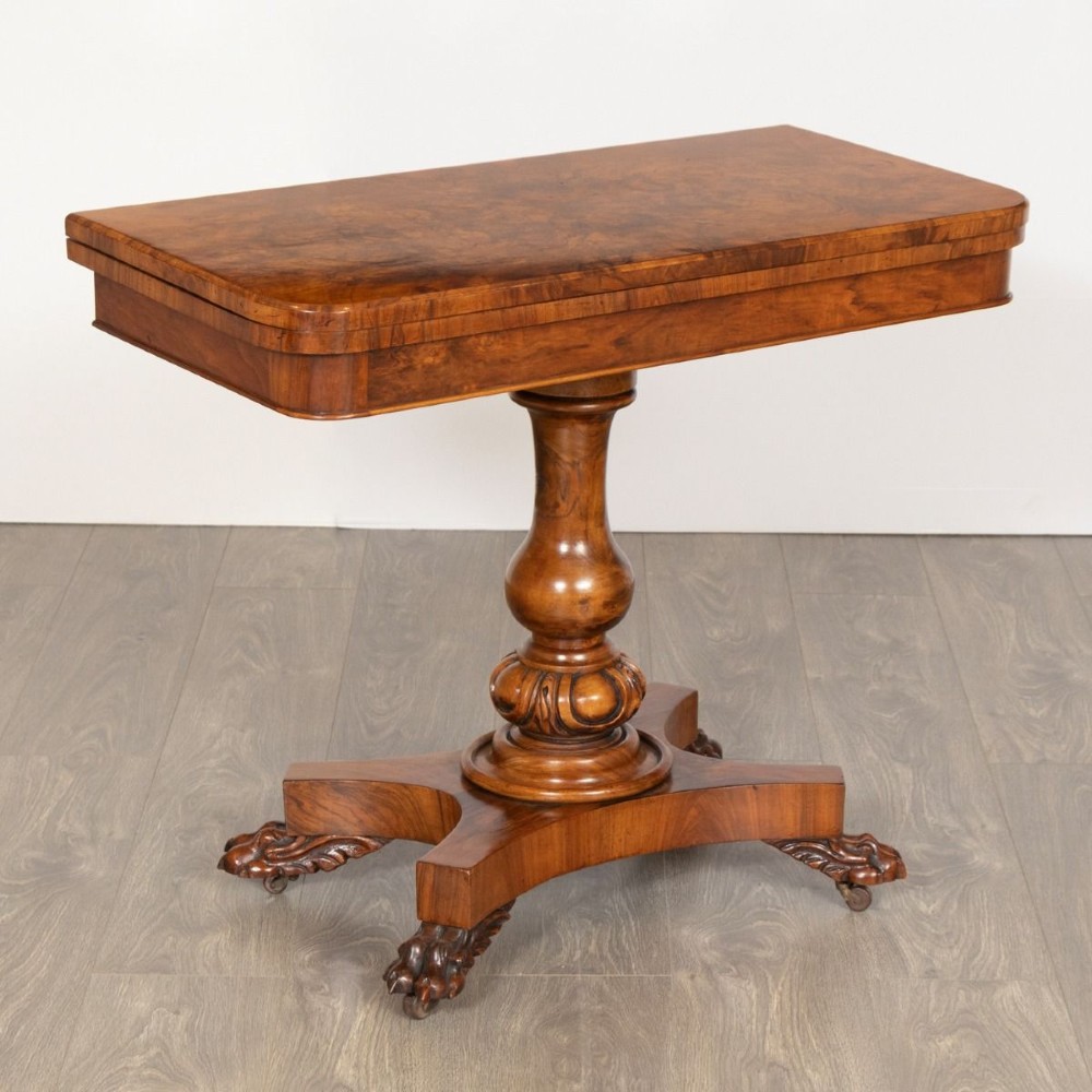 A mid-19th century Victorian burr walnut games table featuring a fold-over rectangular top resting on a turned bulbous pedestal column and a quadripartite base with carved feet on castors. View II