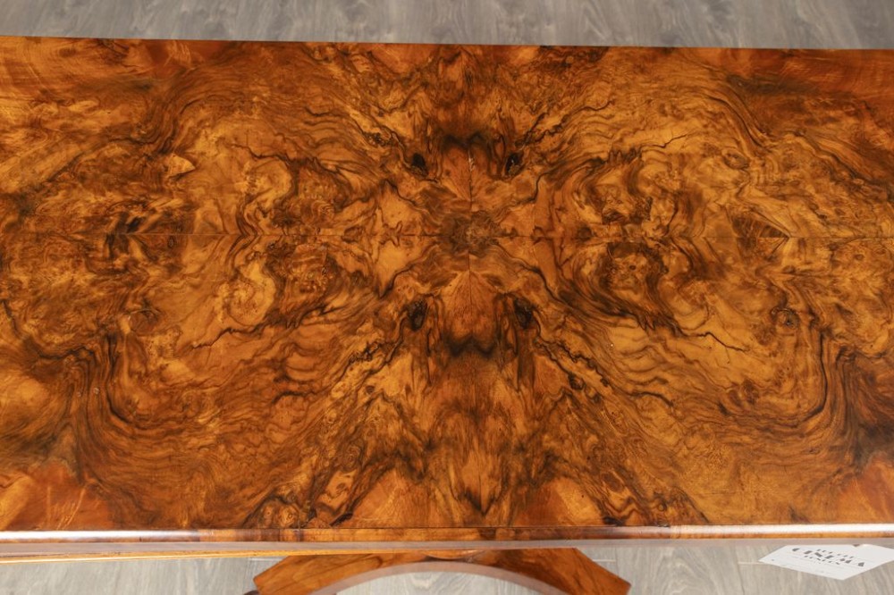 A mid-19th century Victorian burr walnut games table featuring a fold-over rectangular top resting on a turned bulbous pedestal column and a quadripartite base with carved feet on castors. View V