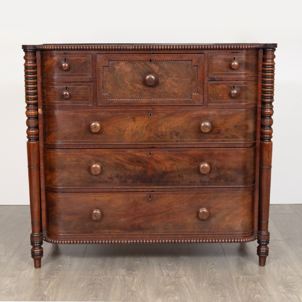 An antique William IV flame mahogany chest of drawers featuring turned spiral columns, multiple small top drawers around a central hat drawer, and wooden turned knobs.