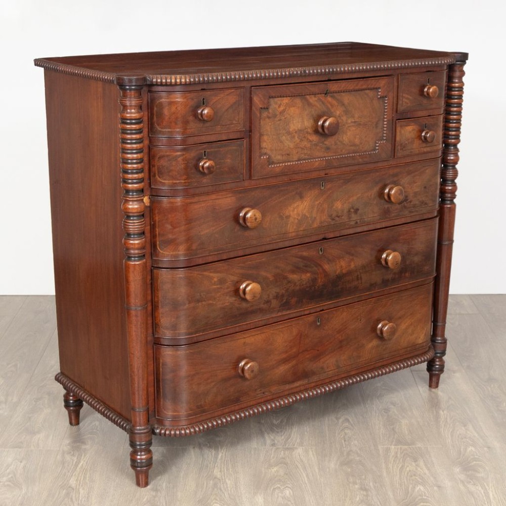 An antique William IV flame mahogany chest of drawers featuring turned spiral columns, multiple small top drawers around a central hat drawer, and wooden turned knobs. View II