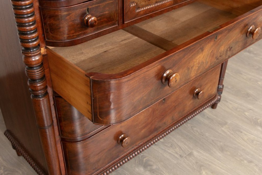 An antique William IV flame mahogany chest of drawers featuring turned spiral columns, multiple small top drawers around a central hat drawer, and wooden turned knobs. View IV