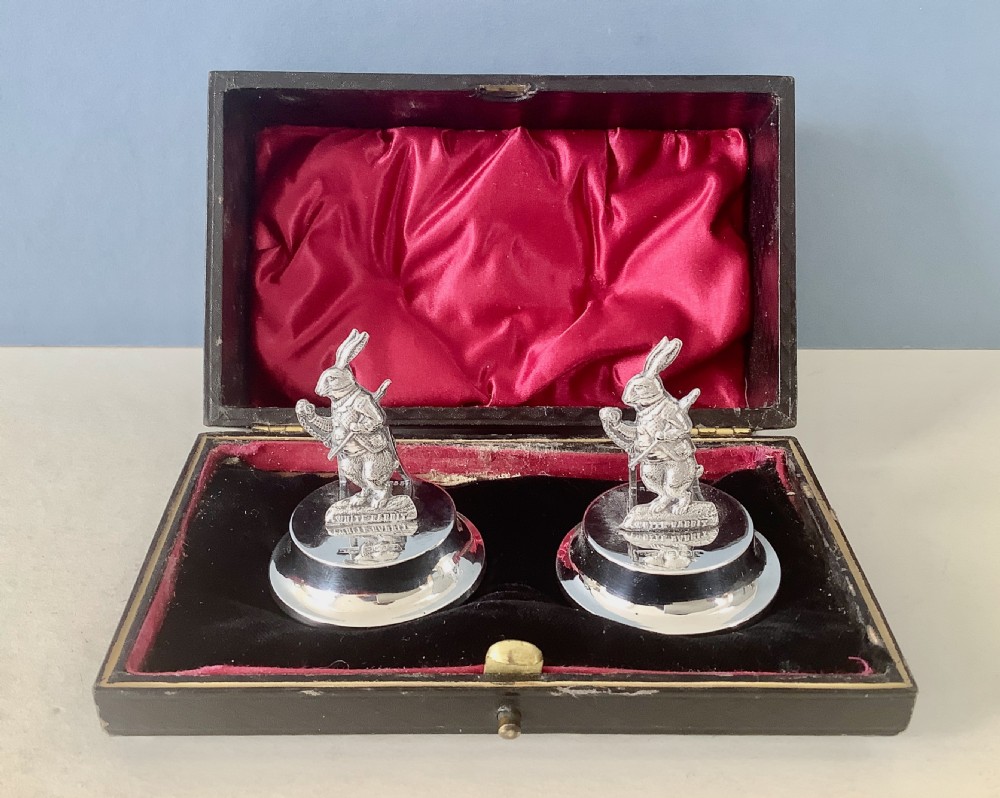 A pair of silver menu holders featuring cast figures of the White Rabbit from Alice in Wonderland, resting on stepped circular bases inside a fitted velvet-lined case.