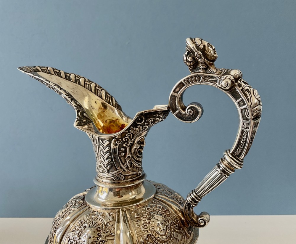 A detailed solid silver Cellini pattern ewer decorated with chased relief masks, scroll motifs, and a sculpted handle atop a pedestal base. View II