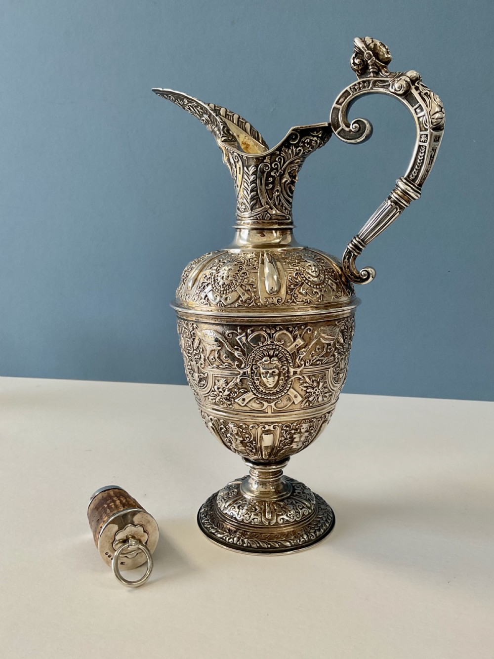 A detailed solid silver Cellini pattern ewer decorated with chased relief masks, scroll motifs, and a sculpted handle atop a pedestal base. View III