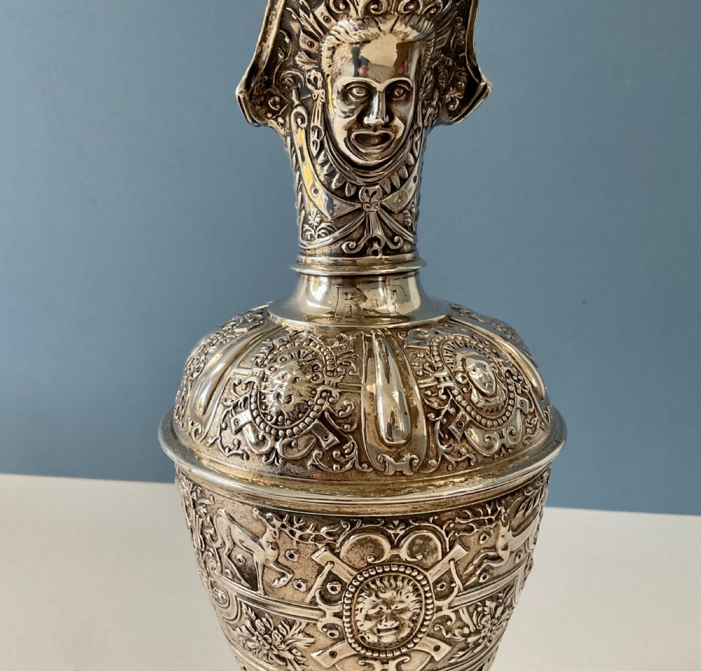 A detailed solid silver Cellini pattern ewer decorated with chased relief masks, scroll motifs, and a sculpted handle atop a pedestal base. View IX