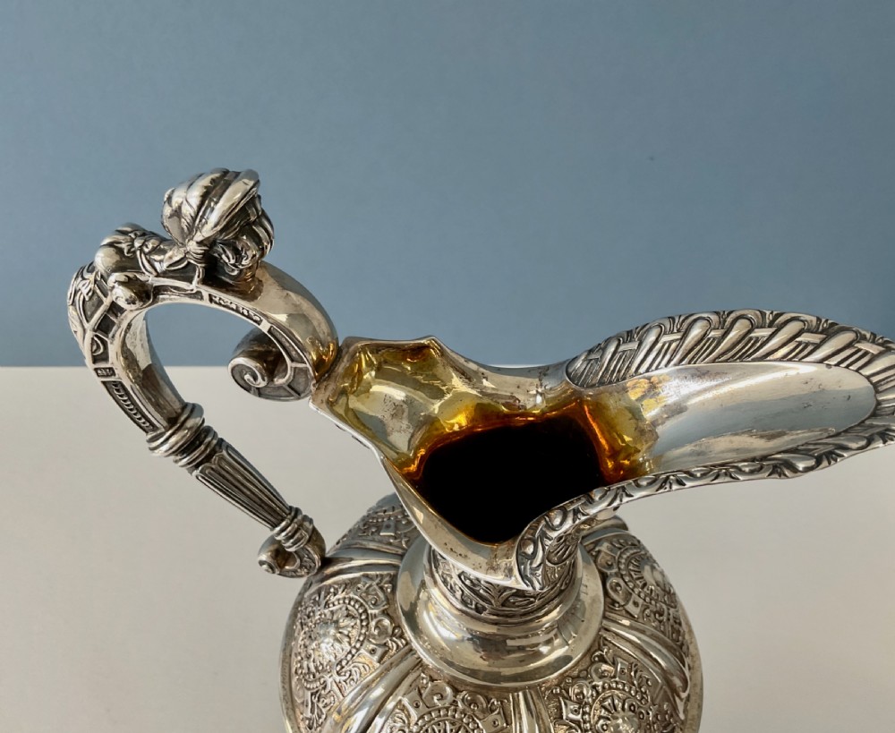 A detailed solid silver Cellini pattern ewer decorated with chased relief masks, scroll motifs, and a sculpted handle atop a pedestal base. View XI