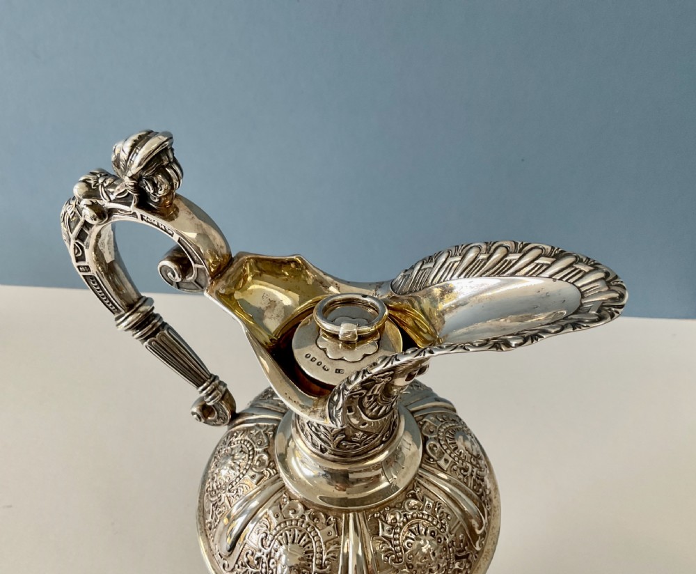 A detailed solid silver Cellini pattern ewer decorated with chased relief masks, scroll motifs, and a sculpted handle atop a pedestal base. View XII