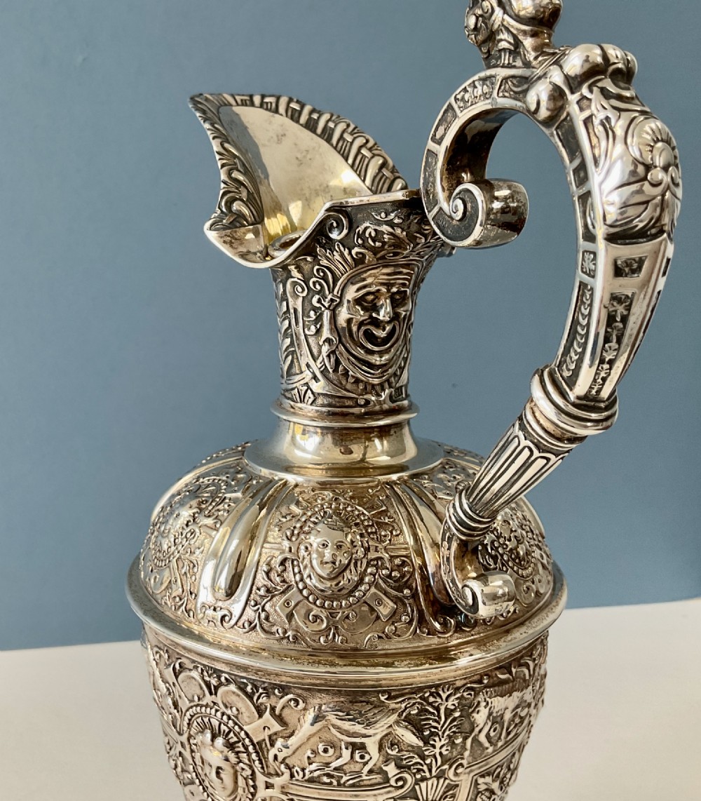 A detailed solid silver Cellini pattern ewer decorated with chased relief masks, scroll motifs, and a sculpted handle atop a pedestal base. View XIV