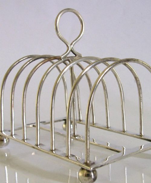 A sterling silver six-slice toast rack featuring seven arched wire divisions, a central loop handle, and four ball feet resting on a rectangular base. View V