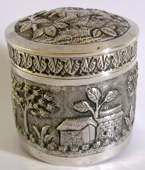 A cylindrical antique Indian solid silver caddy box with a fitted lid, decorated with embossed repousse relief depicting trees, foliage, and village huts. View III