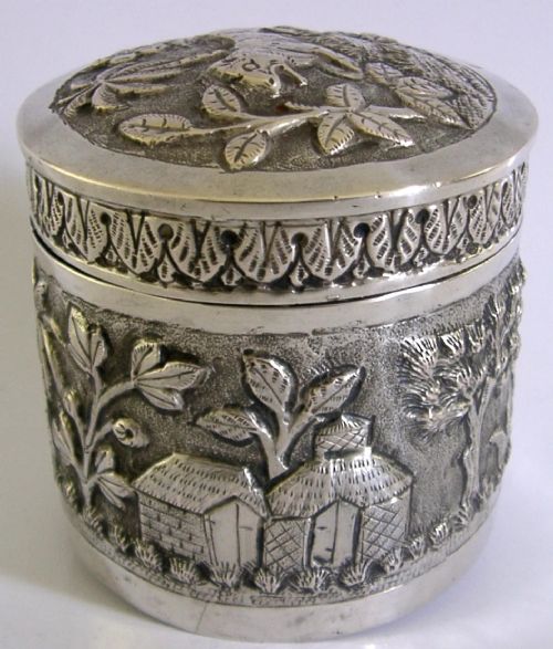 A cylindrical antique Indian solid silver caddy box with a fitted lid, decorated with embossed repousse relief depicting trees, foliage, and village huts. View VI