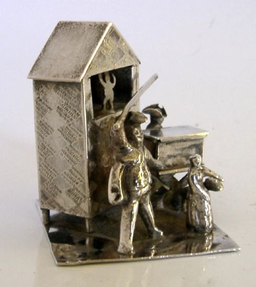 An 833 solid silver miniature model depicting a Punch and Judy puppet show booth with figures, an organ grinder, and a dog on a flat silver base. View II