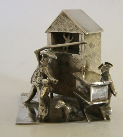 An 833 solid silver miniature model depicting a Punch and Judy puppet show booth with figures, an organ grinder, and a dog on a flat silver base. View III