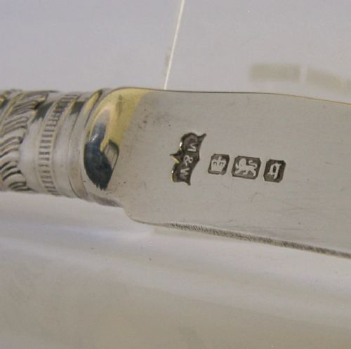 An Edwardian sterling silver cheese knife featuring a curved blade with two terminal prongs, a decorative chased collar, and a smooth cream handle. View V