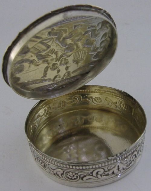 A circular antique silver trinket box featuring embossed relief depicting putti playing beneath floral garlands. View IV