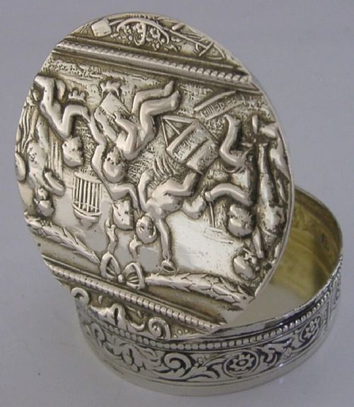 A circular antique silver trinket box featuring embossed relief depicting putti playing beneath floral garlands. View V