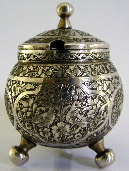 An antique hand-engraved solid silver mustard pot featuring arabesque foliage patterns, a hinged lid with finial, a spoon aperture, and three ball feet. View VI