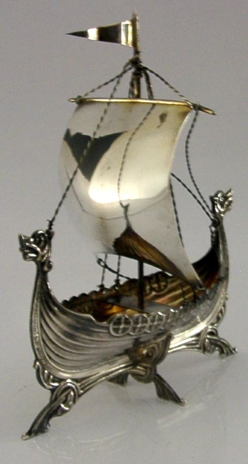 A detailed solid silver model of a Viking longship featuring a dragon head prow, raised sail, pennant flag, and miniature shields along the gunwale. View VI
