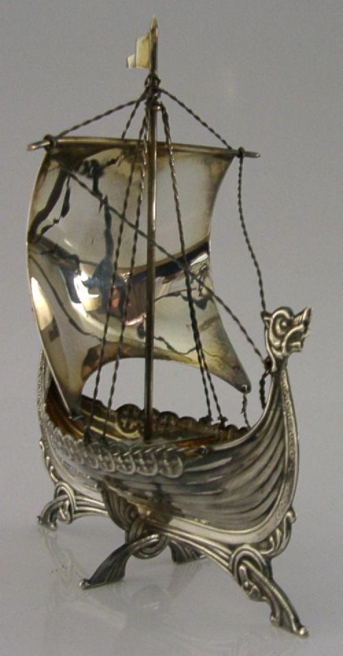 A detailed solid silver model of a Viking longship featuring a dragon head prow, raised sail, pennant flag, and miniature shields along the gunwale. View VII