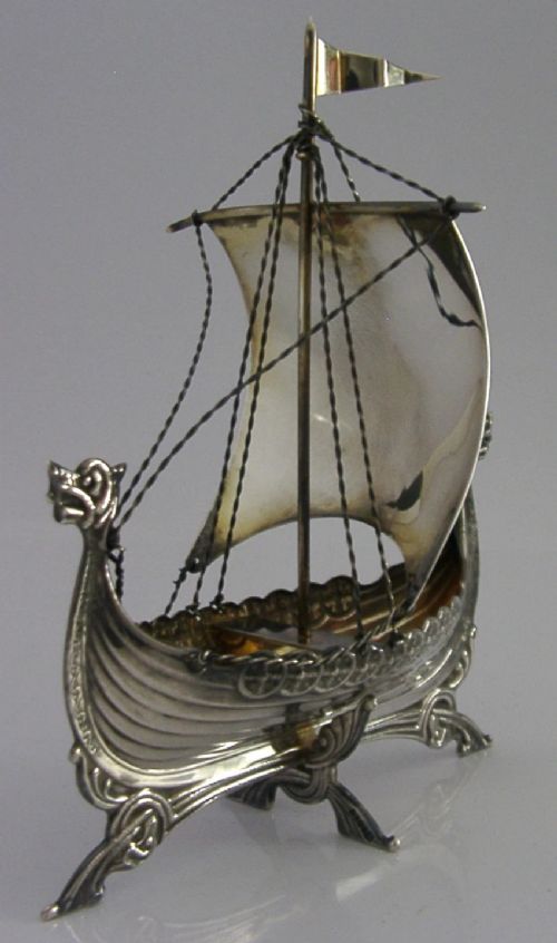 A detailed solid silver model of a Viking longship featuring a dragon head prow, raised sail, pennant flag, and miniature shields along the gunwale. View X