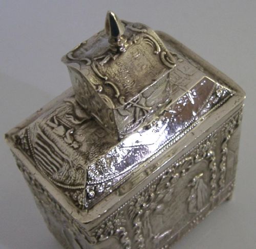 A rectangular solid silver tea caddy featuring relief-embossed pastoral scenes, turned feet, and a finial lid. View VII