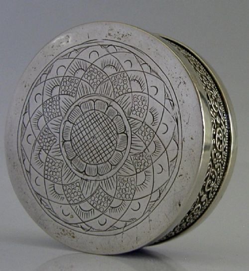 A circular antique silver trinket box with a lidded top featuring dense relief floral scrollwork and an oxidised background. View VI