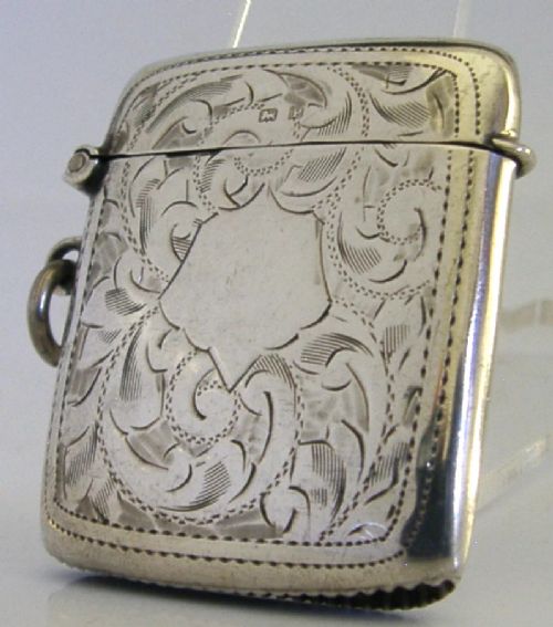 An antique sterling silver vesta case featuring hand-engraved foliate scrollwork, a central shield cartouche, a hinged lid, and a side suspension ring. View VI