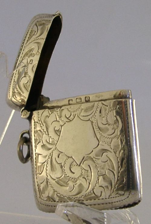 An antique sterling silver vesta case featuring hand-engraved foliate scrollwork, a central shield cartouche, a hinged lid, and a side suspension ring. View VIII