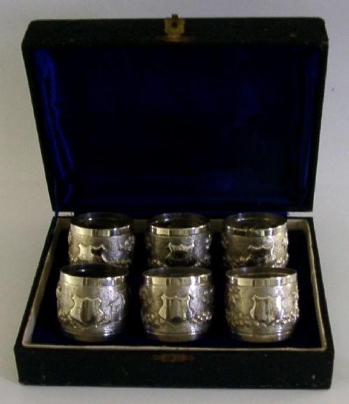 A set of six silver napkin rings with repousse landscape relief patterns and shield cartouches, arranged inside a blue velvet-lined fitted box. View III