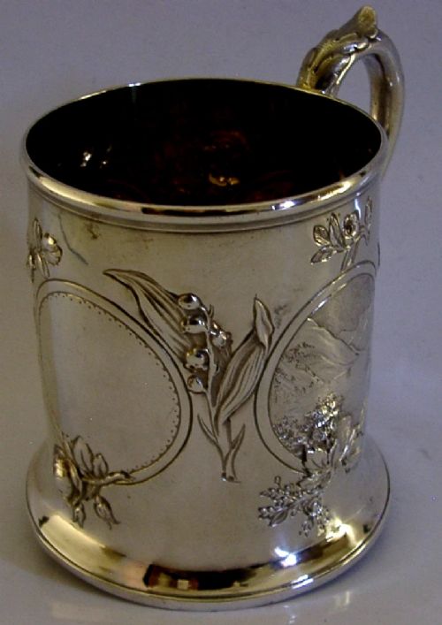 A Victorian sterling silver tankard mug featuring detailed relief floral decoration, a landscape roundel, a scroll handle, and a gilt interior. View VI