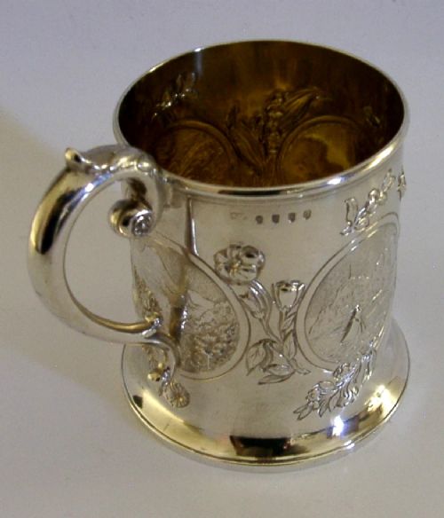 A Victorian sterling silver tankard mug featuring detailed relief floral decoration, a landscape roundel, a scroll handle, and a gilt interior. View VIII