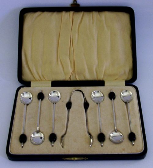 A fitted display case containing six sterling silver coffee spoons with black coffee bean finials and matching sugar tongs. View V